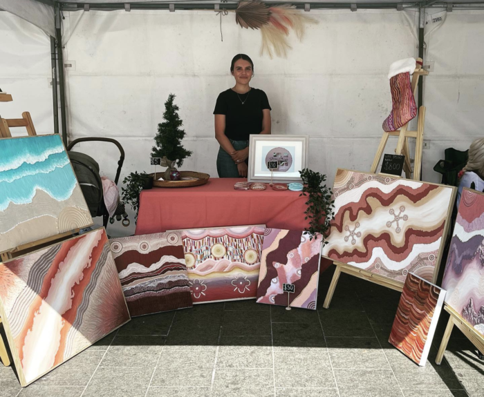A First Nations person stands behind a table at a stand at a market. They are selling paintings.
